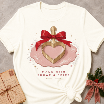 An off-white graphic t-shirt styled flat on a gray textured background, featuring a Valentine's Day themed design with a gold heart-shaped cookie cutter, a red ribbon, and pink icing, with the text 'MADE WITH SUGAR & SPICE'.