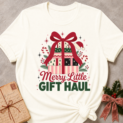 An off-white graphic t-shirt featuring a festive Christmas gift illustration with a large red bow, wrapped presents, candy canes, and the text 'Merry Little GIFT HAUL' in red and green. The t-shirt is styled flat on a textured gray background with a vintage wrapped gift box on the left and holly with a rose gold ribbon on the right.
