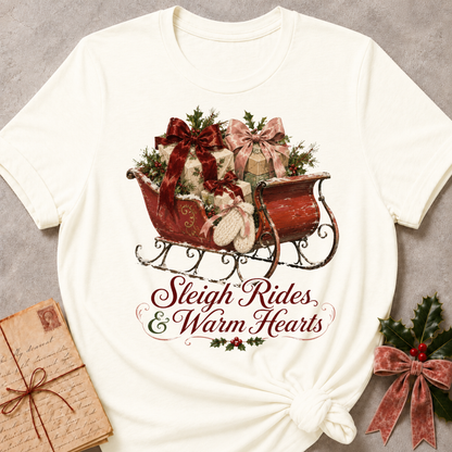 An off-white short-sleeve t-shirt with a Christmas-themed graphic of a red sleigh filled with gifts and greenery, with the text 'Sleigh Rides & Warm Hearts' printed below. The t-shirt is styled flat on a gray textured background with a vintage book and holly in the corners.