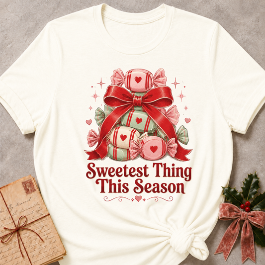 An off-white short-sleeve t-shirt with a Christmas-themed graphic of wrapped candies under a red bow and the text 'Sweetest Thing This Season'. The t-shirt is styled flat on a gray textured background with a vintage book and holly in the corners.