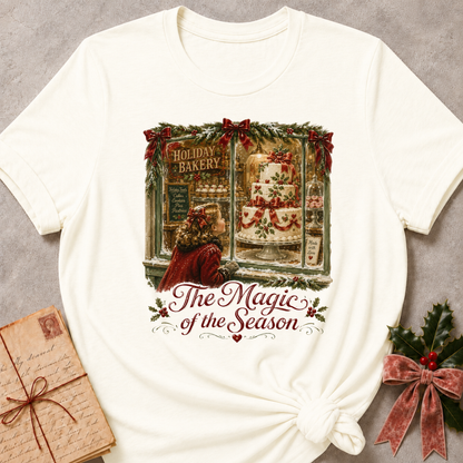 An off-white short-sleeve t-shirt with a Christmas-themed graphic print of a bakery window and the text 'The Magic of the Season'. The t-shirt is styled flat on a gray textured background with a vintage book and holly decoration as props.