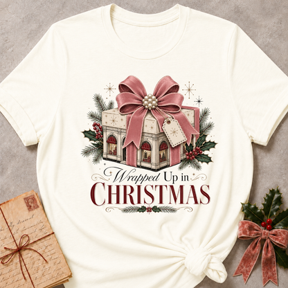 An off-white short-sleeve t-shirt with a Christmas graphic print reading 'Wrapped Up in CHRISTMAS' beneath an illustration of a gift box with a large pink bow, evergreen branches, holly, and red berries. The shirt is styled flat on a gray textured background with a wrapped gift box and holly in the corners.